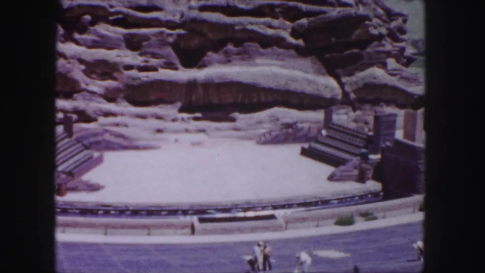Home-movie frame from the 1958 Denver clip showing the rock-lined site, stage edge, stairs, and several tourists near the bottom of frame.