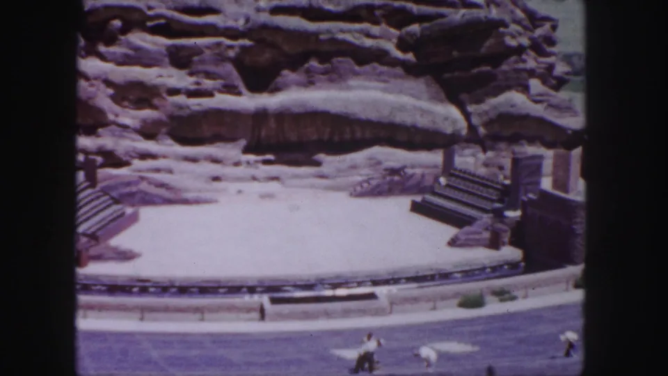 1958 Denver 8mm frame showing distant visitors below a circular stage area, with tiered seating, steps, and textured rock formations behind.