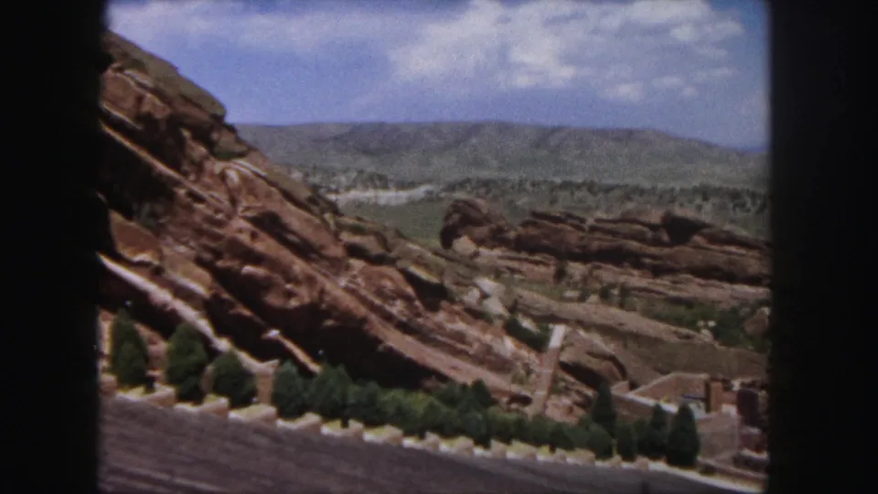 The camera looks across Red Rocks' rocky hillside toward distant Colorado hills under a partly cloudy sky.