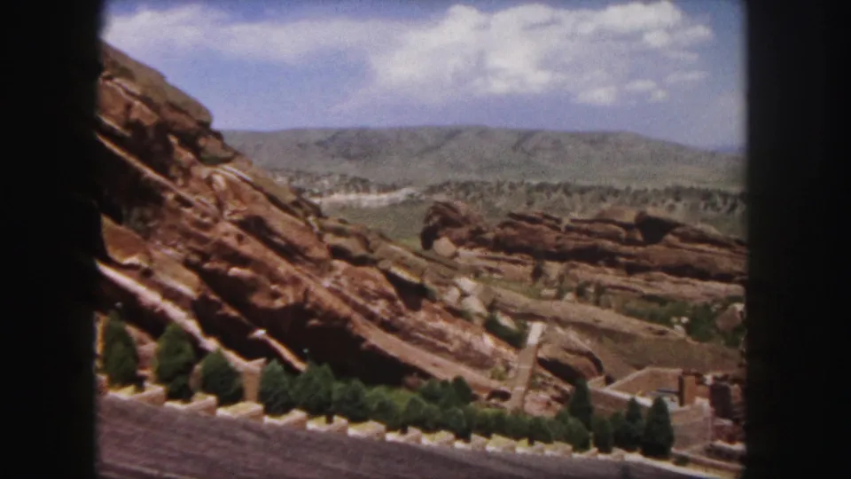 Denver-area 1958 home-movie frame showing layered red rocks, scattered evergreens, distant hills, and bright cloud cover.