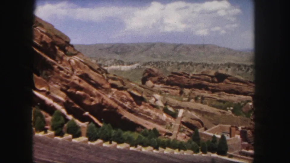 1958 Colorado 8mm still with a red rock foreground, tree line, pale valley, and distant hills under soft clouds.