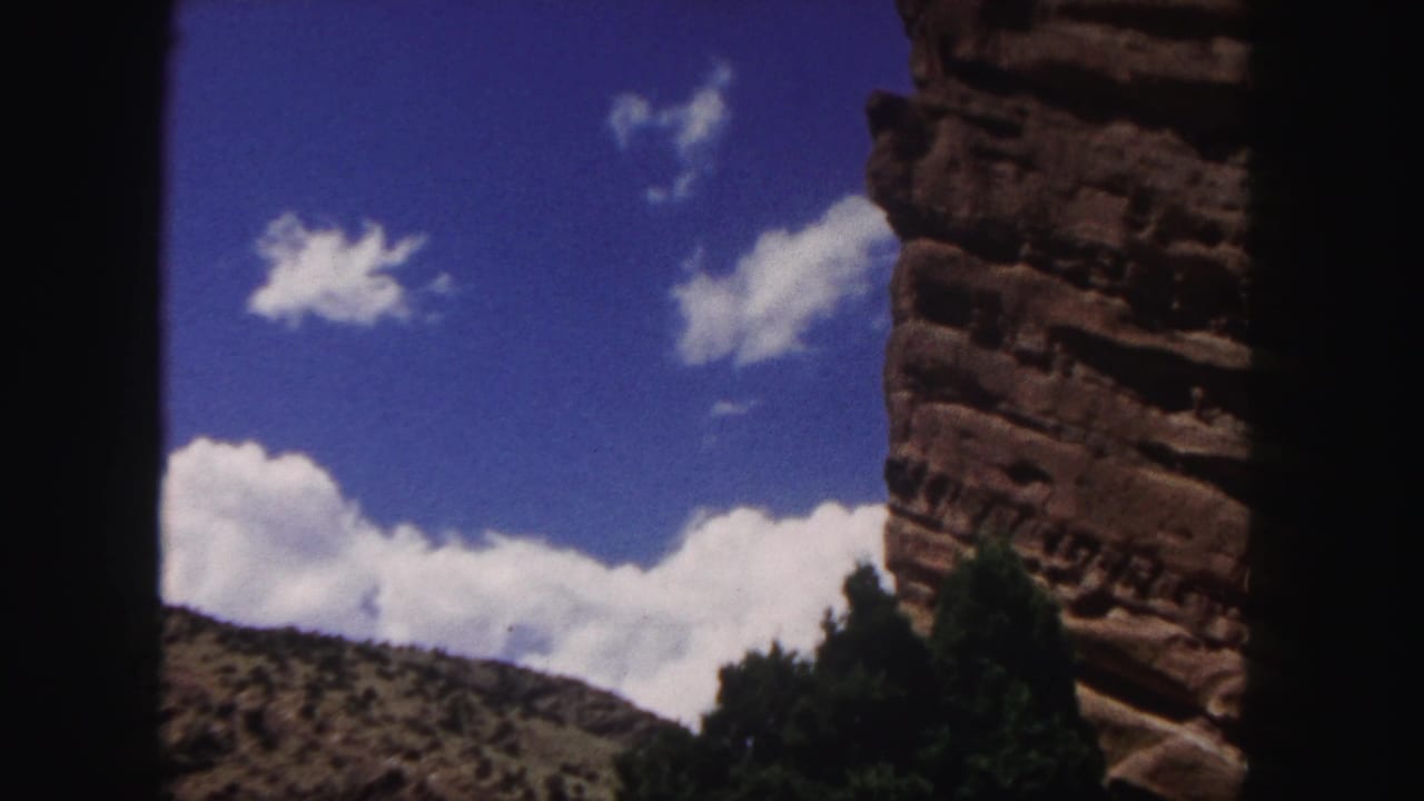 1958 8mm Colorado landscape view with blue sky, white clouds, a red rock cliff at right, distant hillside, trees, and dark frame edges.
