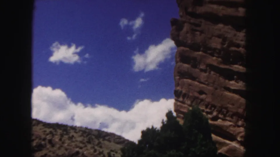 1958 8mm Colorado landscape view with blue sky, white clouds, a red rock cliff at right, distant hillside, trees, and dark frame edges.