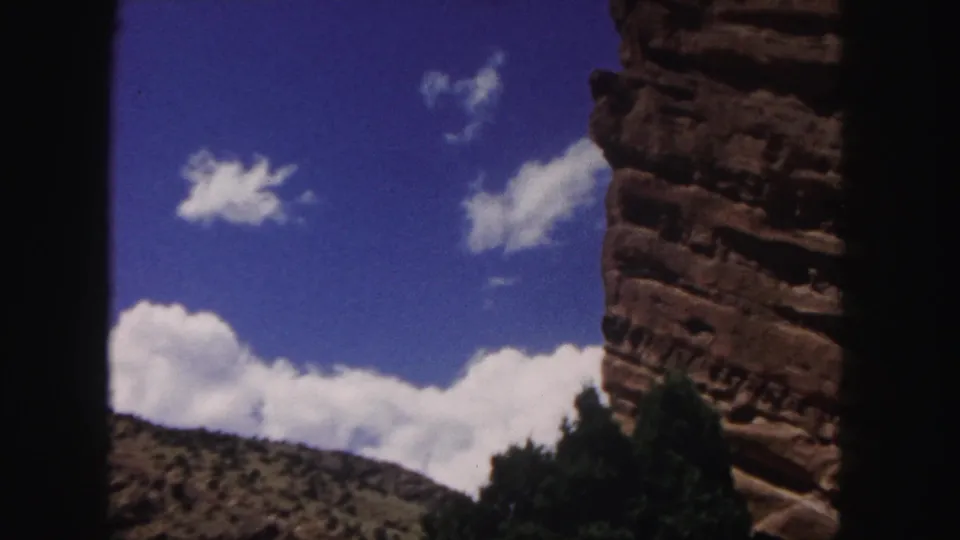 1958 8mm Colorado frame showing a red rock cliff, bright clouds, blue sky, low scrub hillside, trees at the base, and dark scanned film edges.