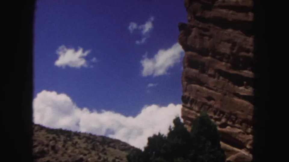 1958 8mm Colorado landscape view with scattered white clouds, blue sky, red rock cliff texture, scrub-covered slope, trees, and visible film-edge framing.