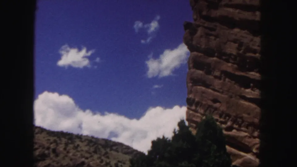 1958 8mm Colorado scenic frame with red rock cliff on the right, blue sky and clouds overhead, green trees below, distant hillside, and dark film-edge shadows.