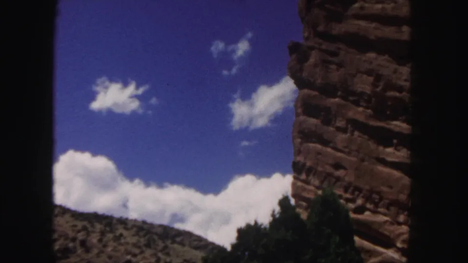1958 Denver-area 8mm frame showing red rock formations, distant hillside, bright clouds, and a dark foreground structure.