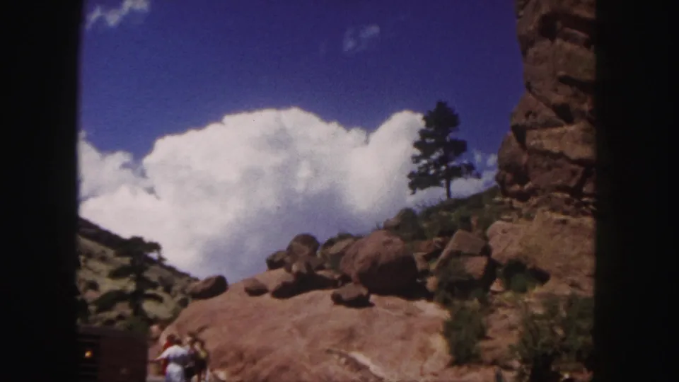 1958 Colorado home-movie frame with distant tourists walking near boulders, a red rock wall, a lone tree, and bright clouds.