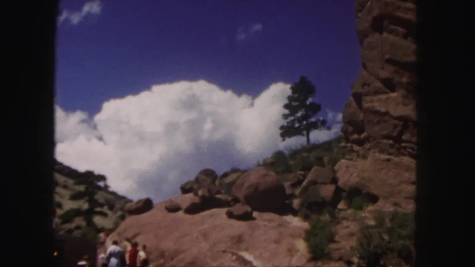 Denver-area 1958 8mm frame showing tourists near rounded boulders, a red cliff edge, a lone tree, and large white clouds.