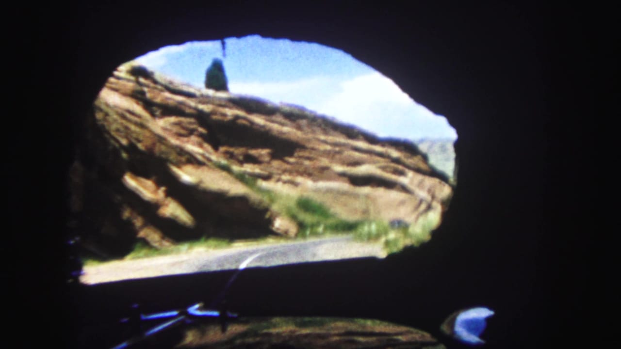 Driver-perspective 8mm frame looking past a tunnel edge toward layered red rock and a curving Colorado road.