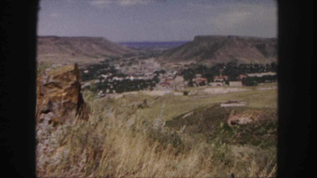 1958 Colorado valley view with town buildings, tree cover, fields, and mesas beyond a rocky overlook.
