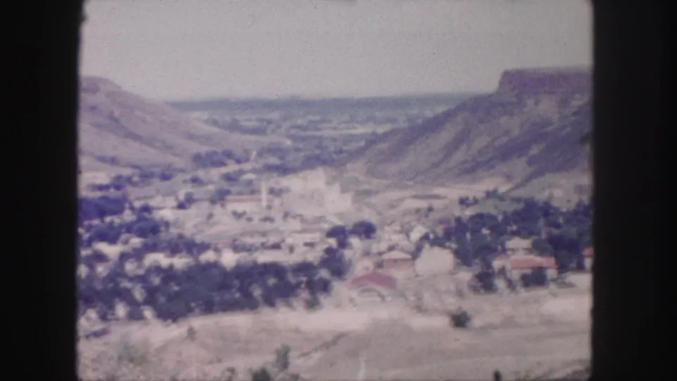 Faded 1958 Colorado frame of a valley town, pale fields, tree cover, and distant rugged bluffs.