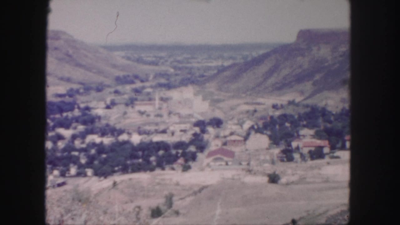 1958 8mm view near Denver showing a small valley settlement, dry hills, and a flat-topped ridge under a pale sky.