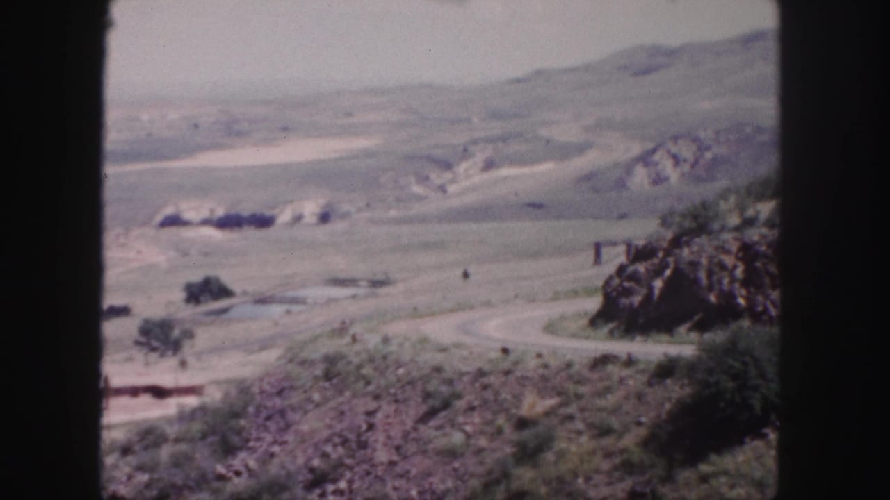 1958 Colorado home-movie frame looking across a dry valley, winding road, rocky foreground, and distant hills near Denver.