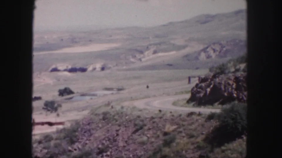 1958 Colorado home-movie frame looking across a dry valley, winding road, rocky foreground, and distant hills near Denver.