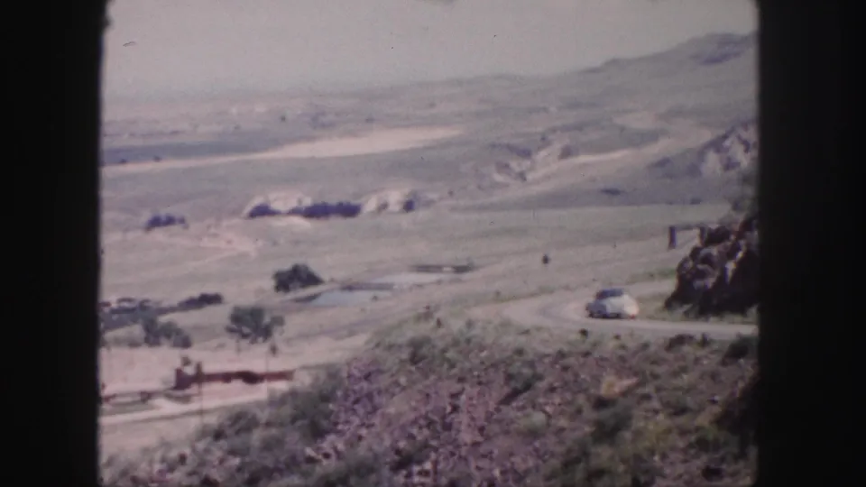1958 8mm Colorado road scene with a white car traveling along a curve through open hills near Denver.