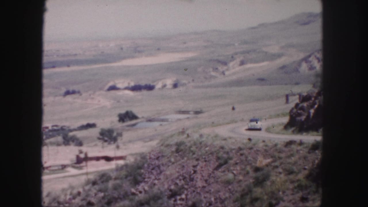 1958 8mm view near Denver showing a winding road across an arid hillside landscape with scattered trees and distant terrain.