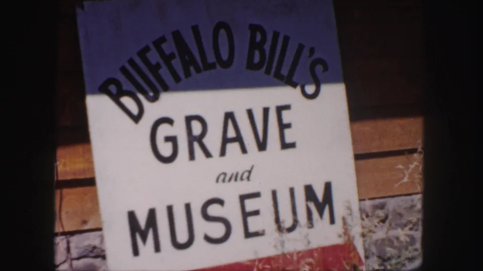 Close 1958 8mm view of the Buffalo Bill's Grave and Museum sign with wood siding, stonework, and dry grass nearby.