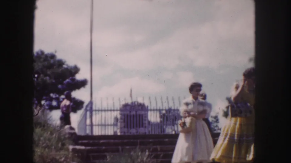 1958 Denver home-movie frame showing two women in mid-century dresses near a metal fence, trees, steps, and cloudy sky at the grave site area.