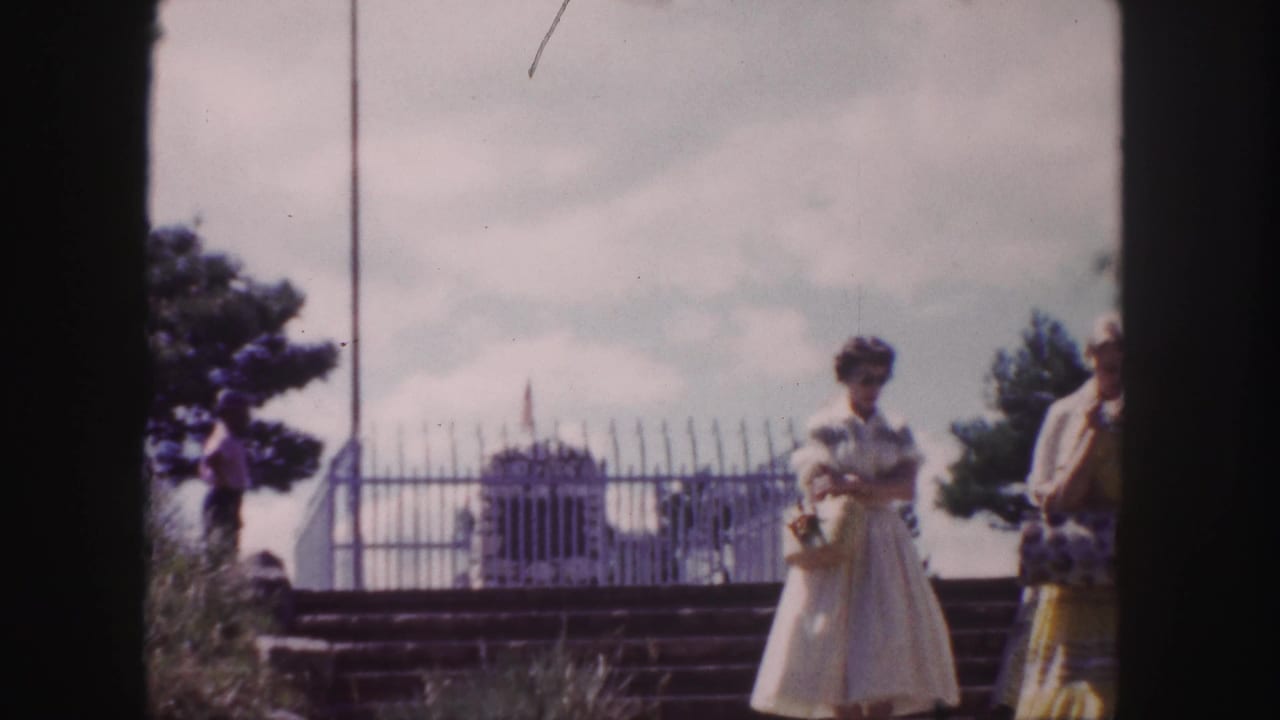 1958 Denver home-movie frame showing visitors near a fenced monument, with a flagpole, trees, and cloudy sky in the background.