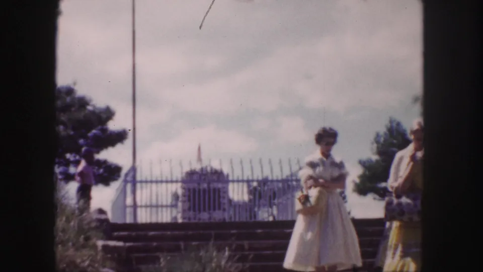 1958 Denver home-movie frame showing visitors near a fenced monument, with a flagpole, trees, and cloudy sky in the background.