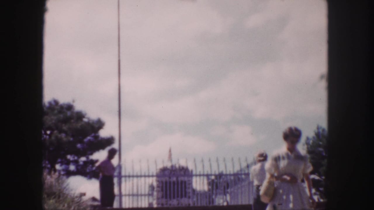 Several visitors walk past a metal fence with a monument and flagpole behind them.