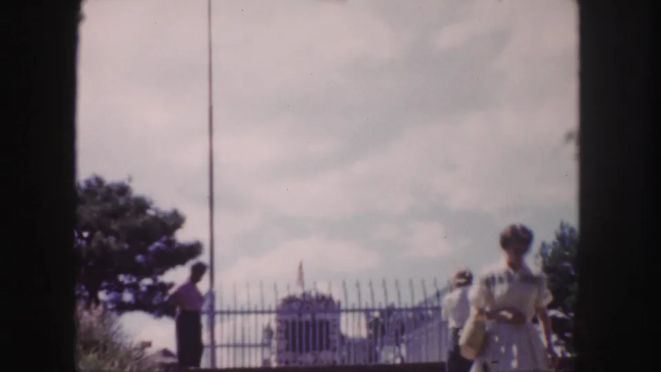 1958 Denver frame with tourists moving along a path beside a metal fence, monument, flagpole, and trees.