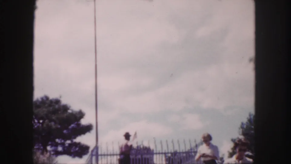 1958 Denver home-movie view of a hat-wearing man by the fenced monument, with trees and sky behind him.
