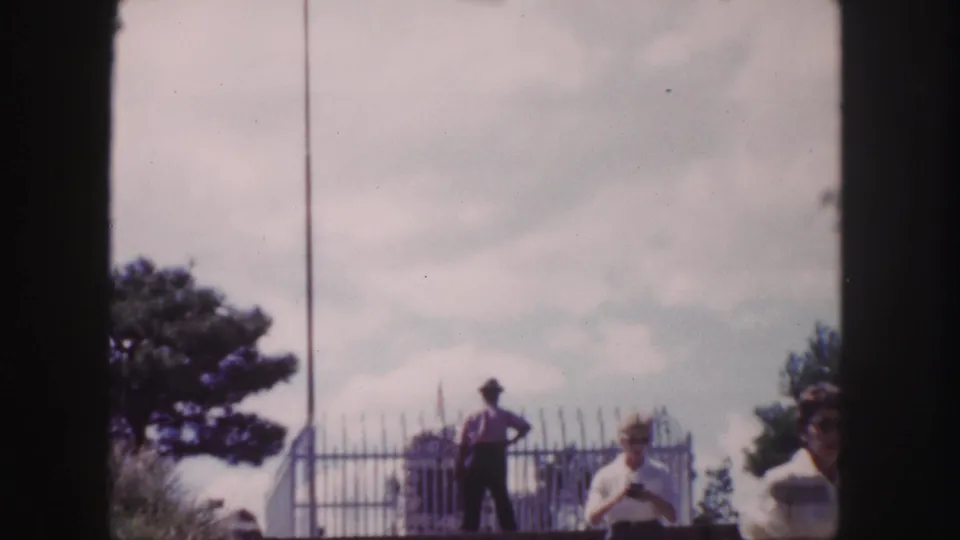 1958 Denver frame showing visitors near a metal fence and monument, including a silhouetted man and a woman in light clothing.