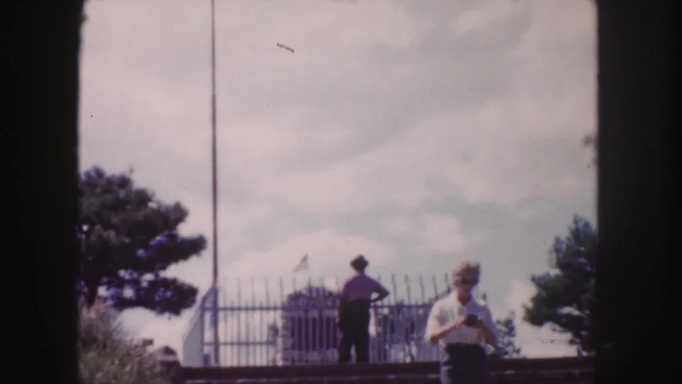 1958 Denver home-movie frame showing two adults near an ornate metal fence with trees, sky, and distant landscape visible.