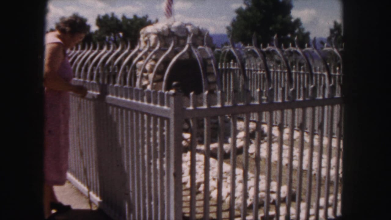 1958 Denver frame with a woman in a pink dress beside an ornate fence around a stone monument or marker.