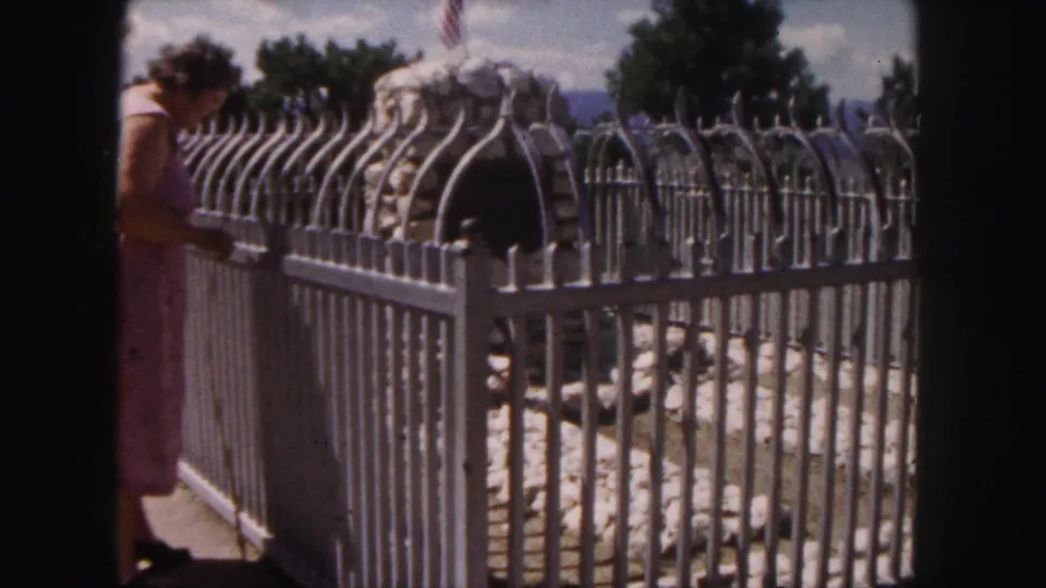 1958 Denver frame with a woman in a pink dress beside an ornate fence around a stone monument or marker.