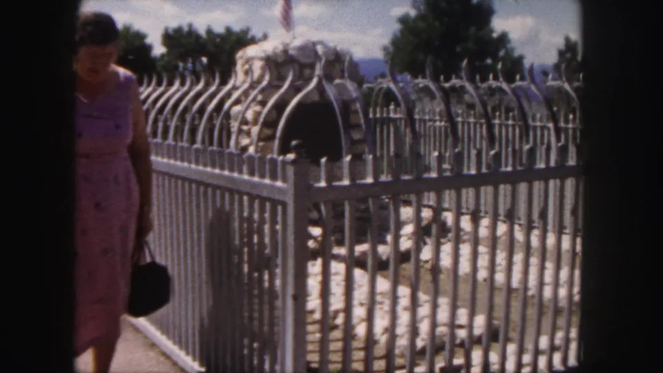 1958 Denver home-movie still of a woman walking beside the ornate fence around stonework, with trees and sky behind.