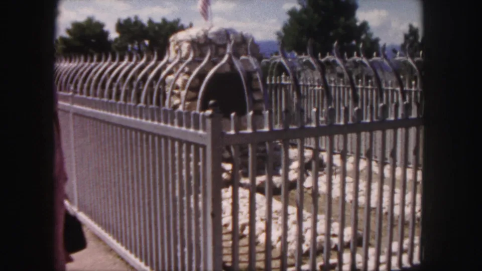 1958 Denver frame focused on the ornate metal fence and stone monument, with the woman partly visible at the edge.