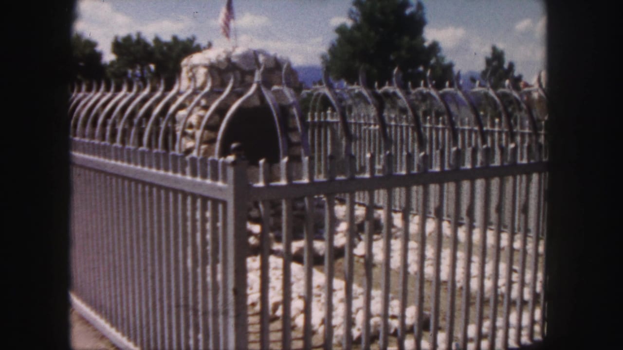 Second 1958 Colorado frame repeats the fenced stone feature, with vertical metal bars, trees, and a flag visible beyond the rocks.