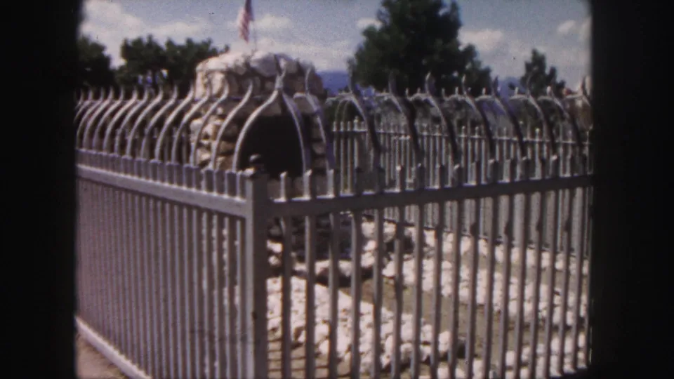 Second 1958 Colorado frame repeats the fenced stone feature, with vertical metal bars, trees, and a flag visible beyond the rocks.