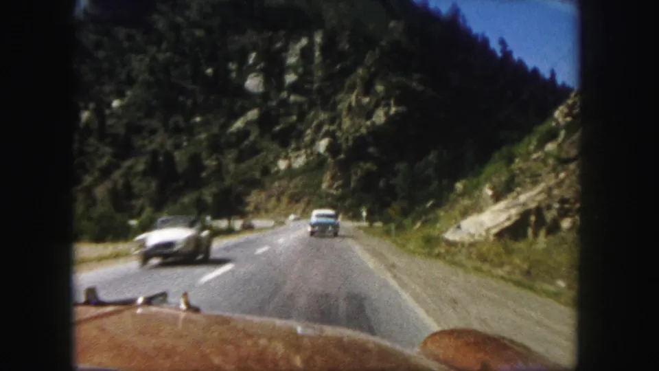 1958 Colorado road-trip frame showing a mountain highway filmed from a moving car, with oncoming vehicles, rocky slopes, and forested hillsides.