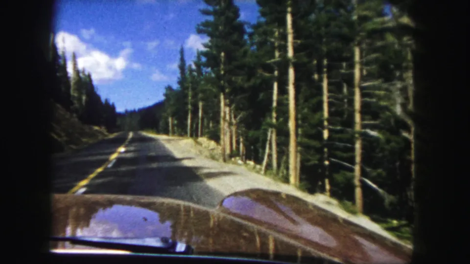 1958 8mm Denver-area driving view with a car hood, paved mountain road, pine forest, blue sky, and clouds.