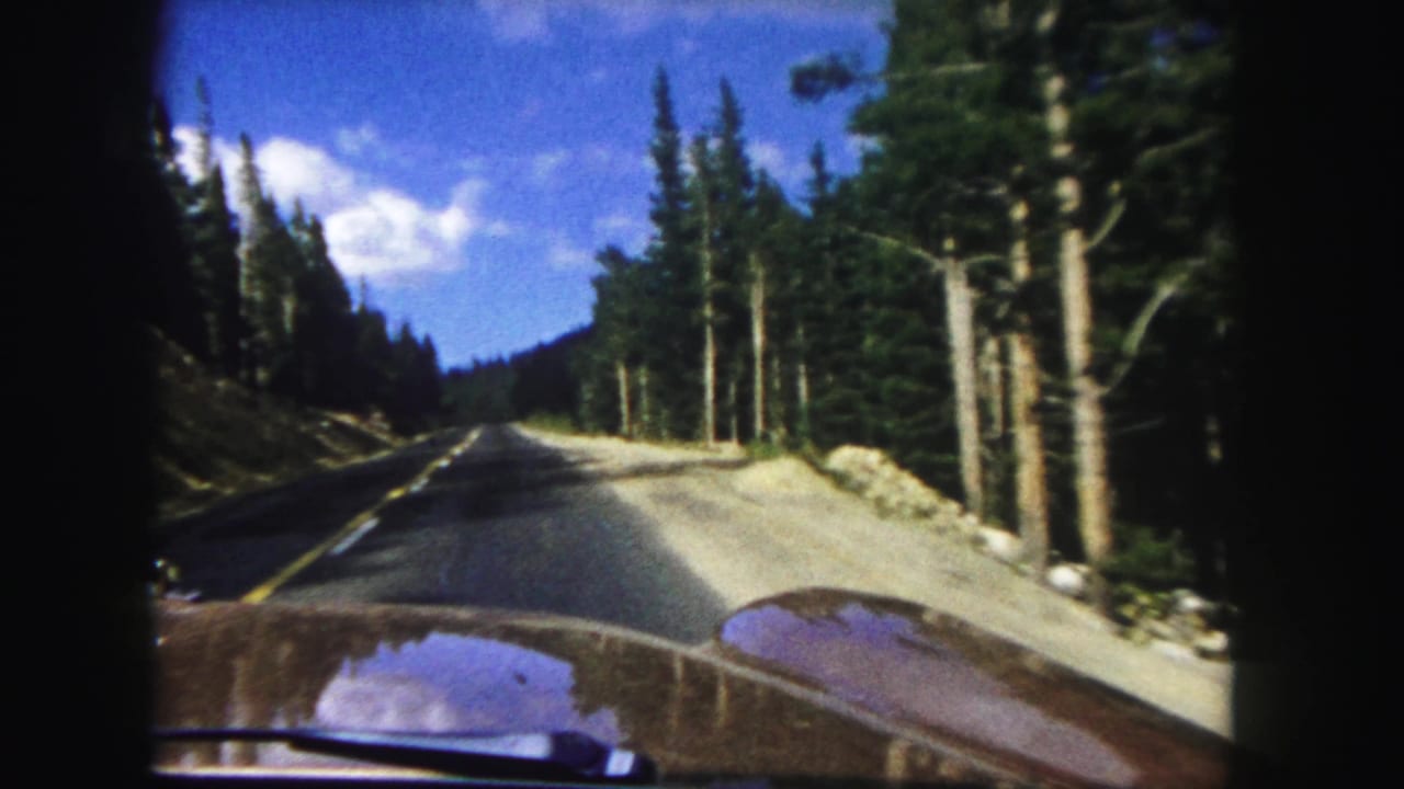 Denver-area 1958 home-movie frame looking over the car hood toward a pine-lined mountain road.