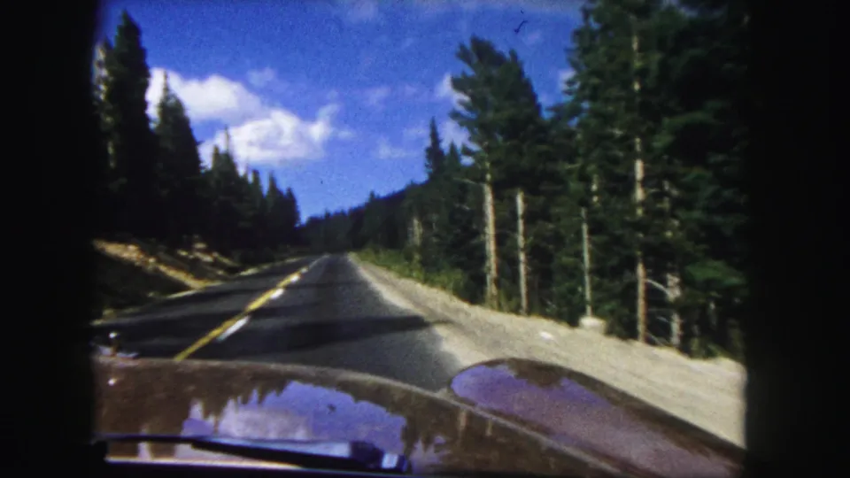 Denver-area 1958 8mm frame of a car traveling along a pine-lined mountain road under blue sky.