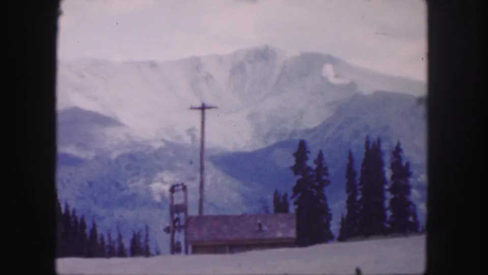 1958 8mm view of a snowy Colorado mountain ridge with dark evergreens, a lodge roof, a utility pole, and a ski-lift tower in the foreground.