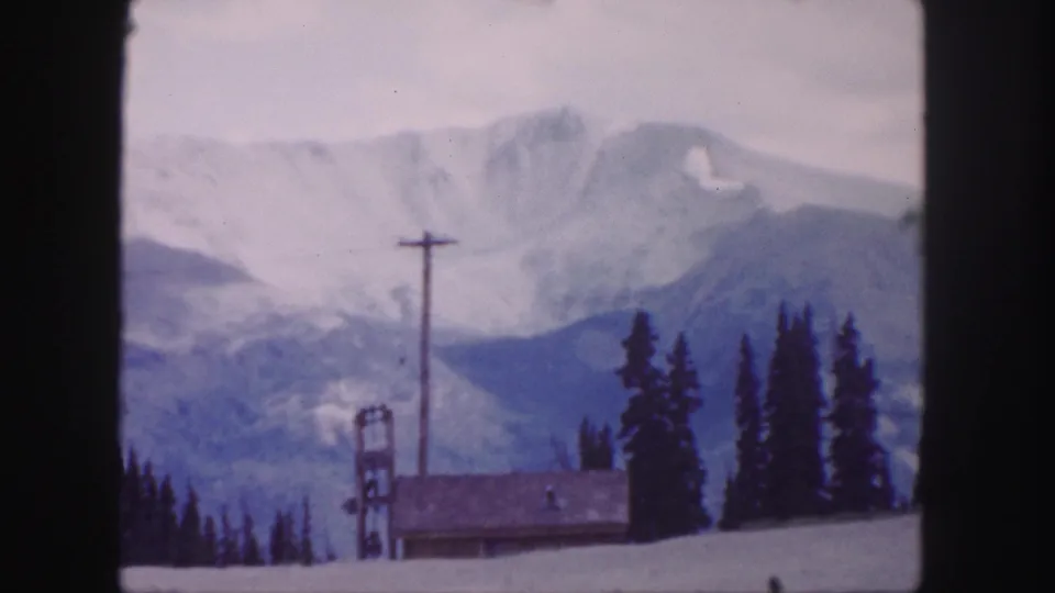 1958 Colorado home-movie frame showing a soft snowy mountain view near Berthoud Pass, with evergreens, lodge roof, utility pole, and lift tower.