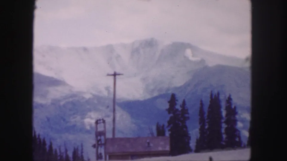 1958 Colorado home-movie frame with a cabin below snowy mountain ridges, a central utility pole, and evergreen trees.