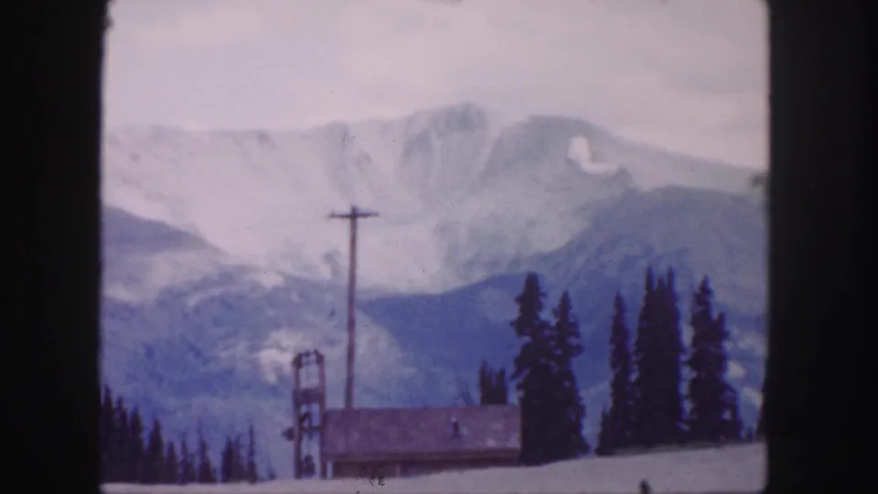 1958 Colorado 8mm view of a mountain cabin and utility pole framed by evergreens and hazy snow-covered peaks.