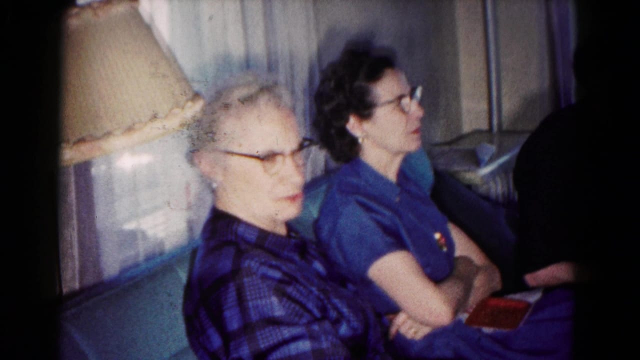 In a 1958 Hagerstown home interior, two women wearing glasses sit closely on a sofa beside a table lamp and curtains.