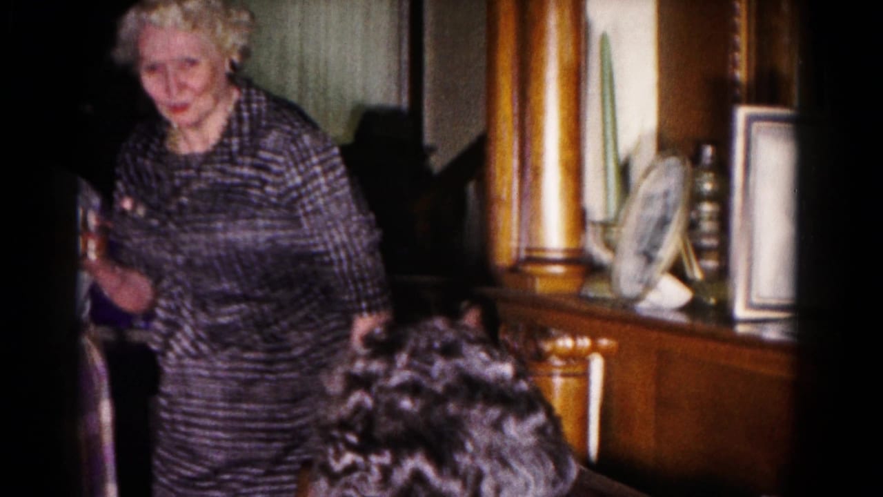 Older woman in a patterned dress standing or seated near wood furniture in a living room.