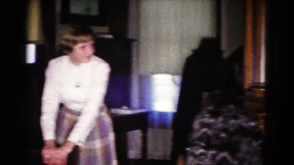 Young female family member in a white blouse and plaid skirt sits in the 1958 Hagerstown living room near a lamp and dark wood furniture.