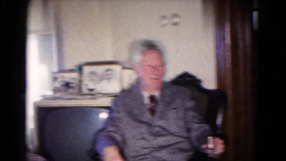 Older man in a gray suit sits beside a boxy television set and framed objects in the 1958 Hagerstown family living room.