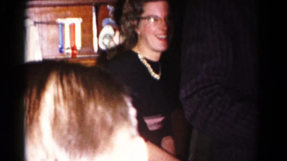 A smiling woman with glasses sits near shelves as other adults stand close together in the 1958 Hagerstown home interior.