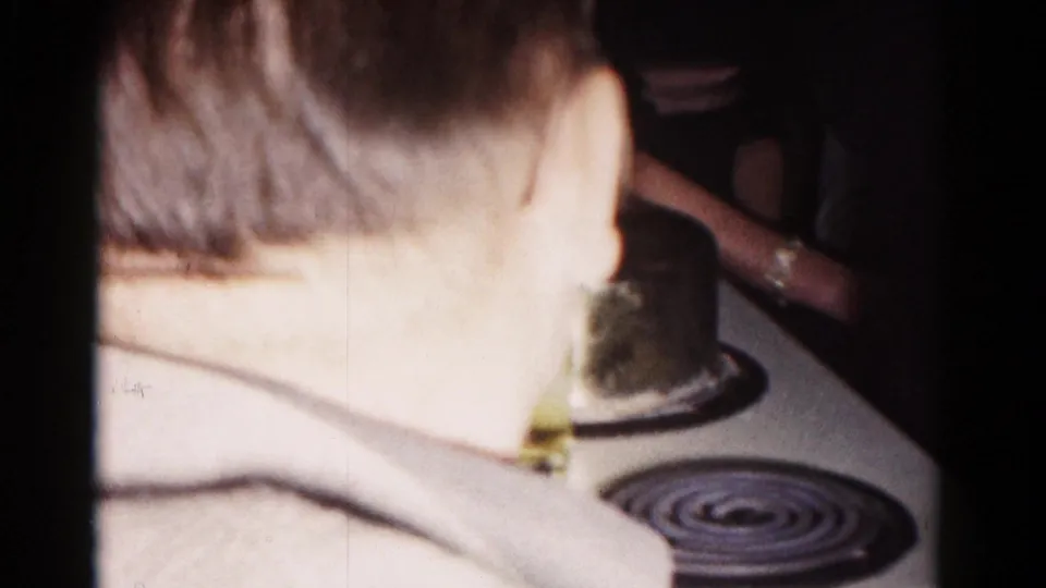 In a 1958 Hagerstown home-movie frame, a man is seen from behind beside a kitchen stove as others stand nearby in dim light.